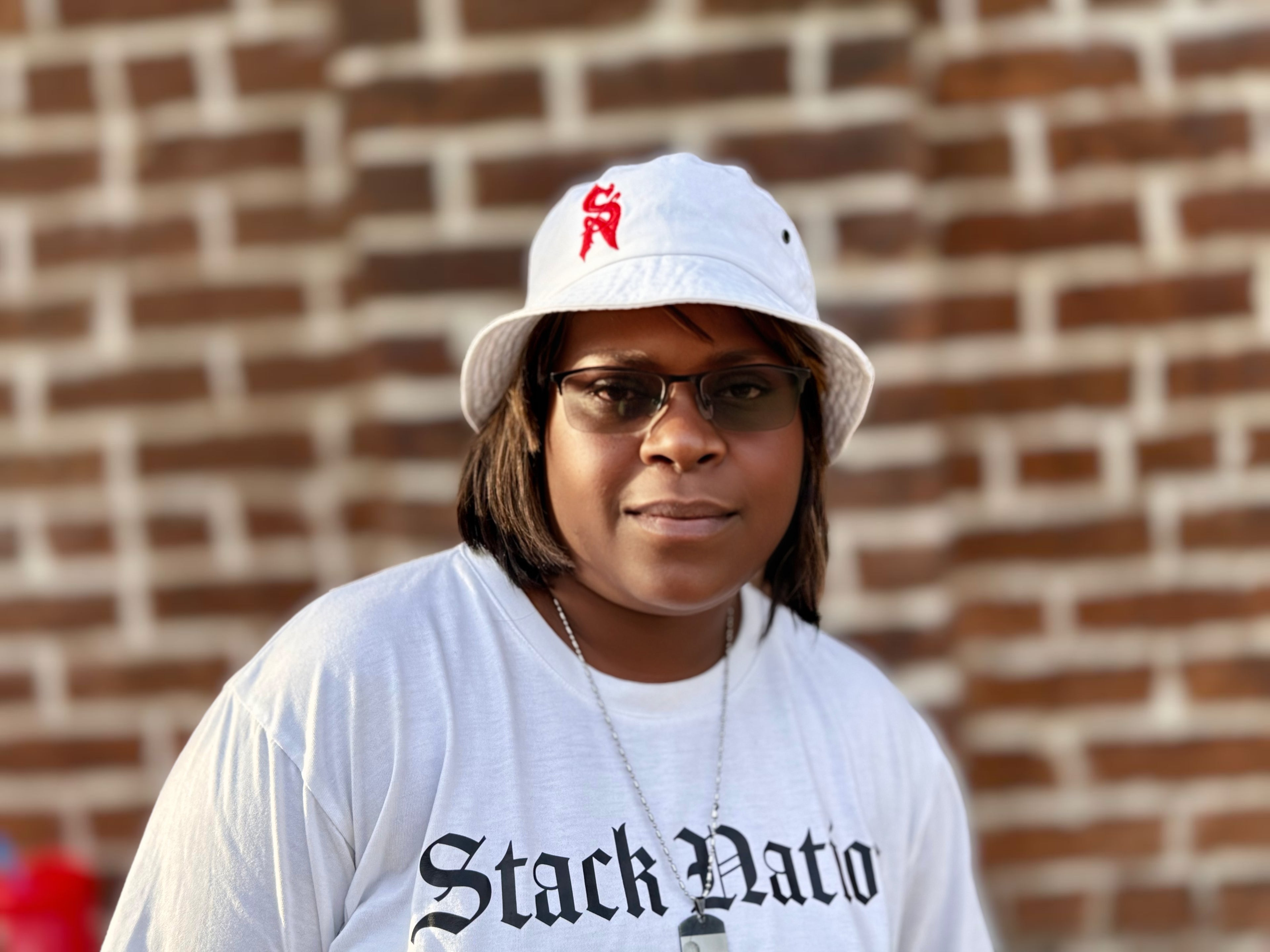 Person wearing a STACK NATION LLC, white cap and t-shirt with text, standing against a brick wall.