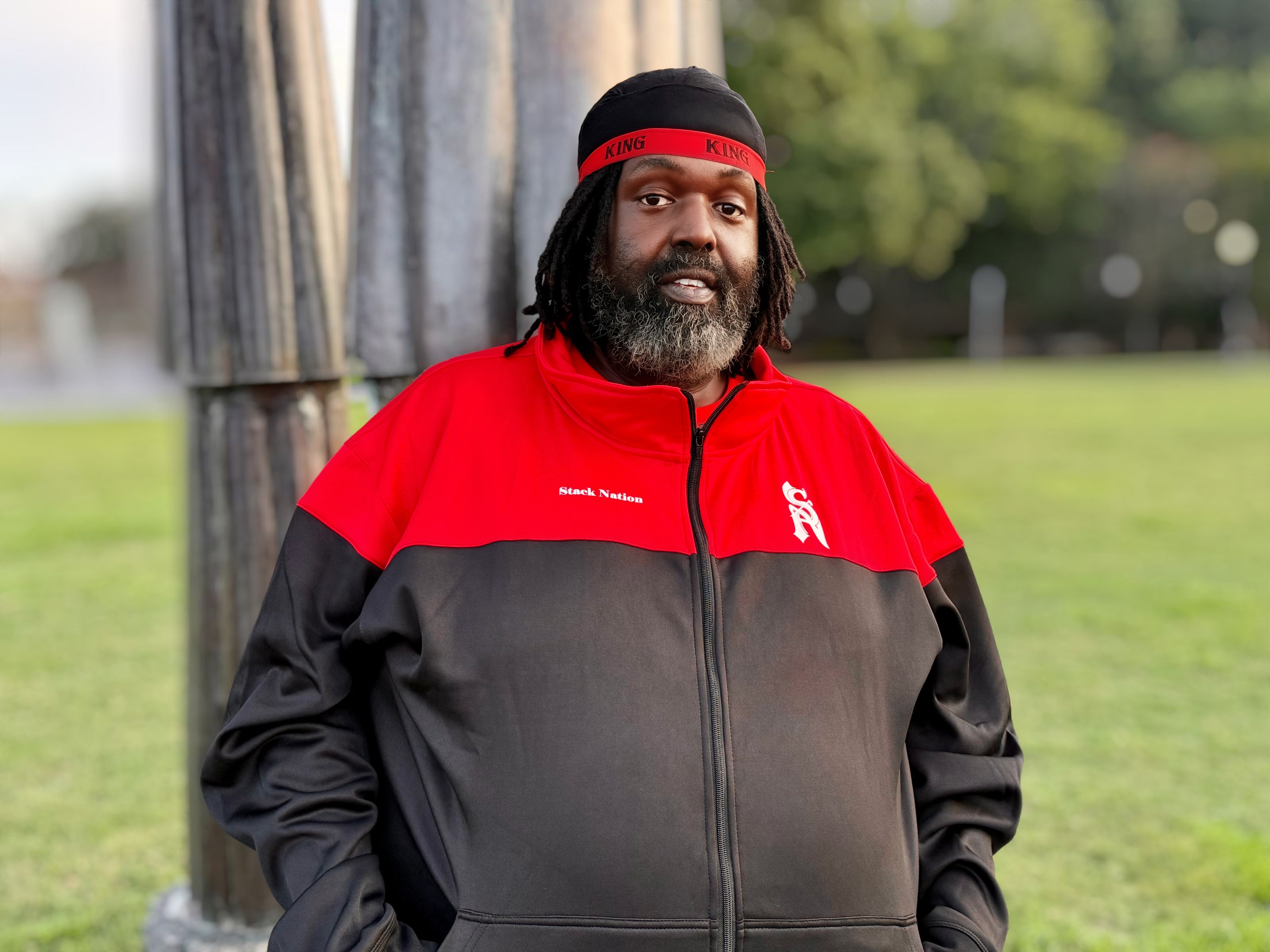 Plus Size Man wearing a black and red jacket with a logo, standing outdoors on grass.