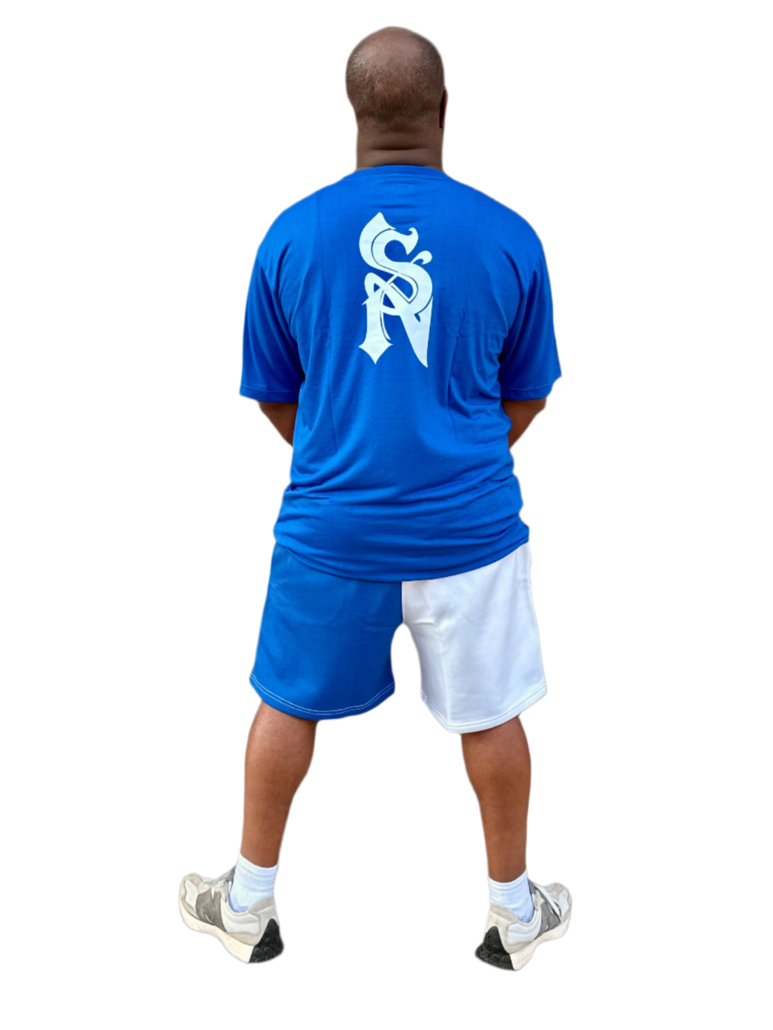 Person wearing a blue t-shirt with a logo and shorts, standing against a brick wall.