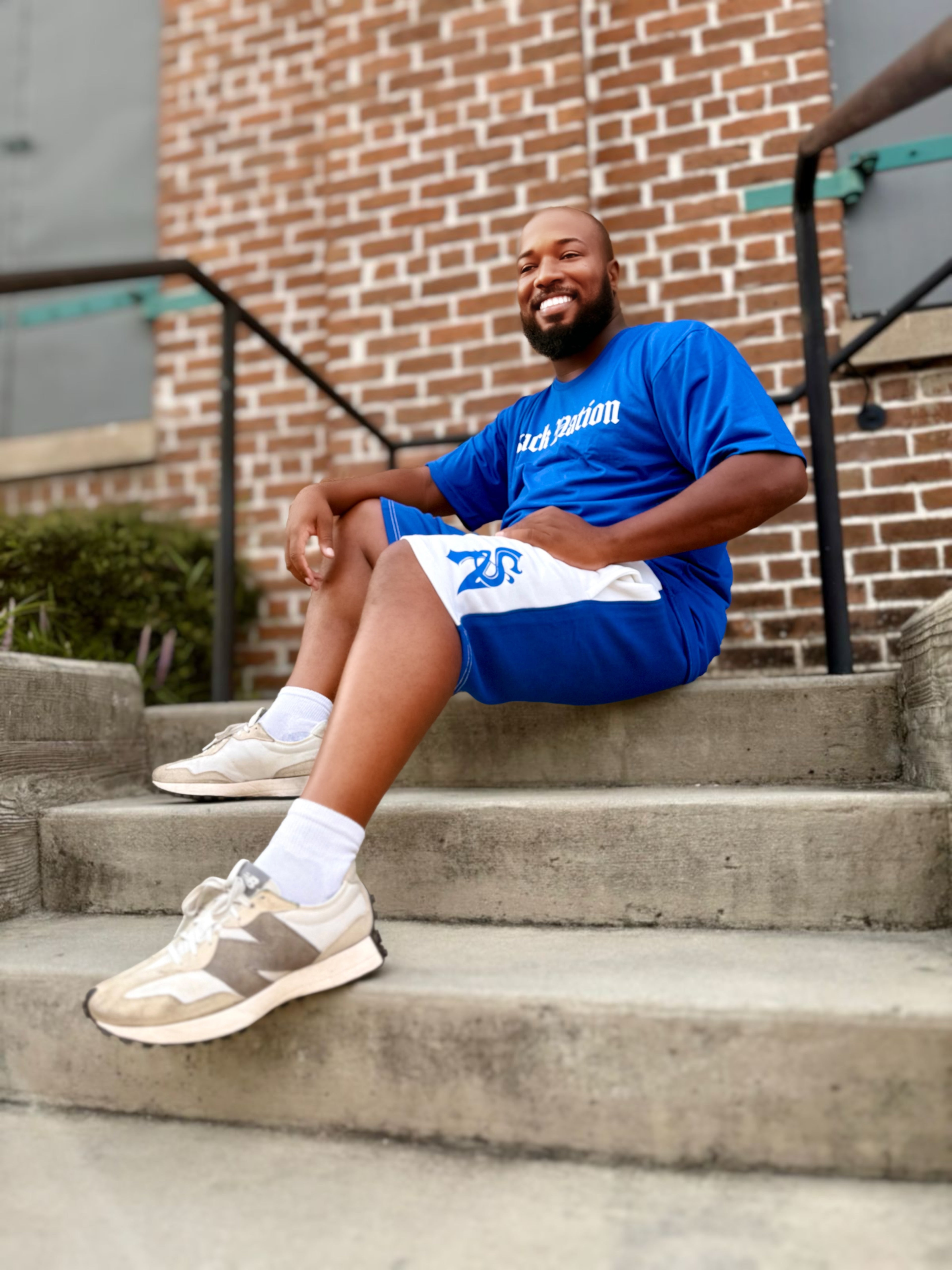 Man sitting on steps wearing a blue t-shirt and shorts with visible branding.